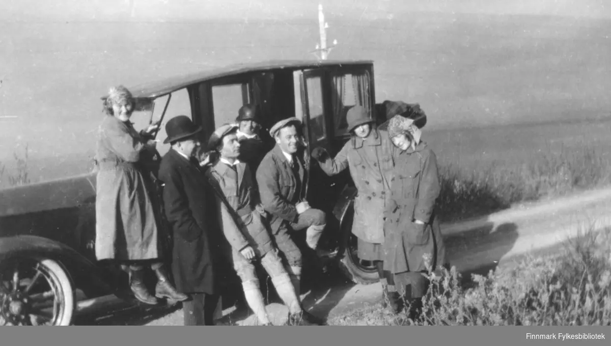 Fotografi av fire kvinner og tre menn som står ved en bil som er er fra 1910- eller 1920-tallet. Damenes klokkehatter er typisk 20-talls. De er alle kledt i fritidsklær utenom en mann som er kledt i en mørk frakk og hatt. Bilen står på en grusvei. 