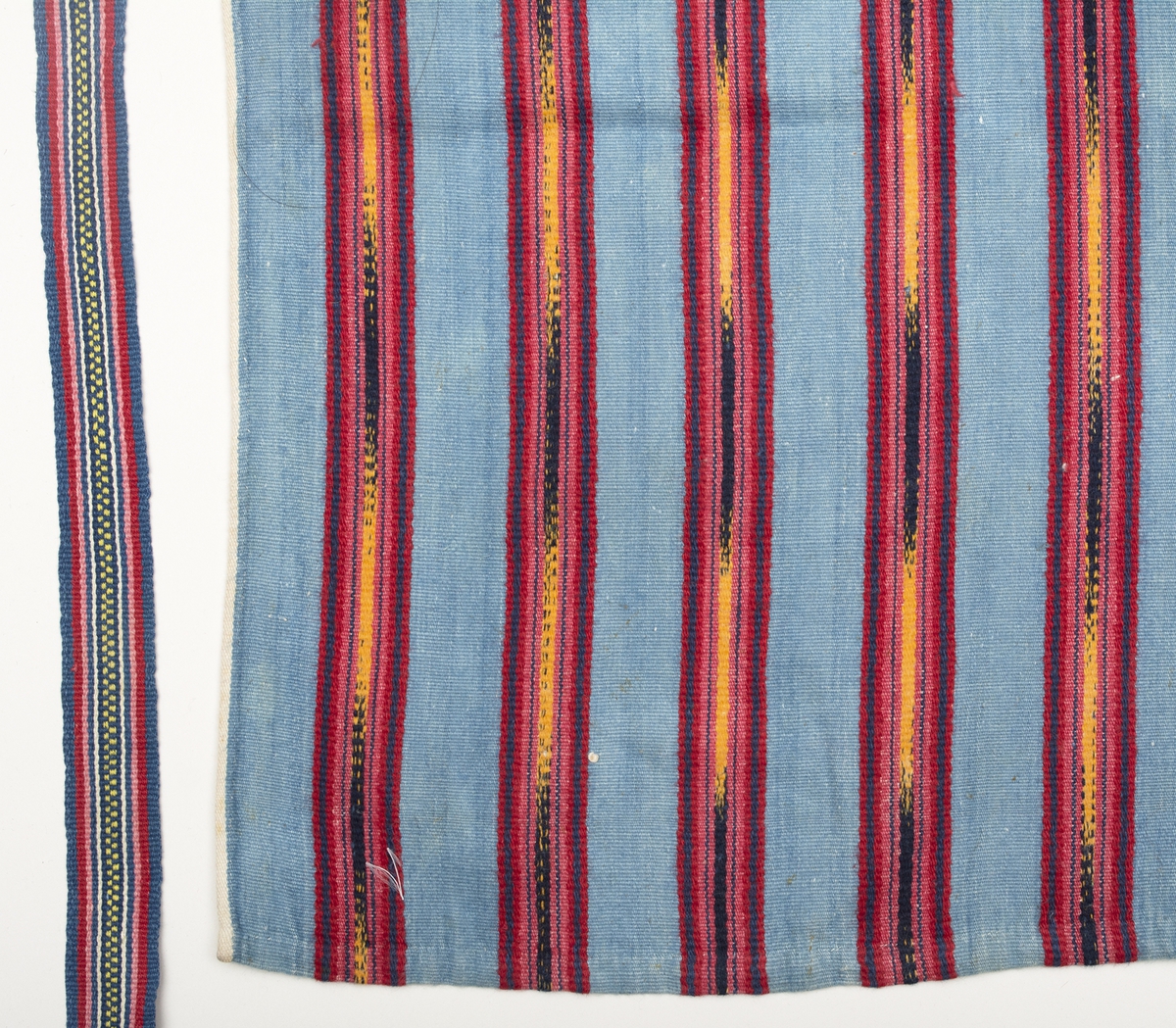 Midjeförkläde med ljusblå botten och breda jämt fördelade ränder i rött, gult och mörkblått, där de gula och mörkblå är flamgarnsfärgat. Ytterst längs långsidorna löper en smal vit kant som fortsätter på baksidan i den 4 mm breda sidfållen. Fåll nertill ca 10 mm.
Linning av ett tyg liknande förklädets, bredd 15 mm, dubbelvikt över förklädeskanten. I sidorna är 1150 mm långa handvävda, mönstrade knytband fastsydda.