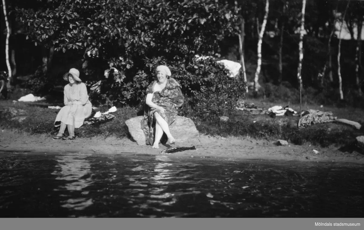 Fru Ester Alberts (1883 - 1968, gift med Karl Alberts) sitter på en sten vid Tulebosjön, cirka 1930. Mattor brukade tvättas på stenen då vattnet var högre. Okänd dam till vänster.