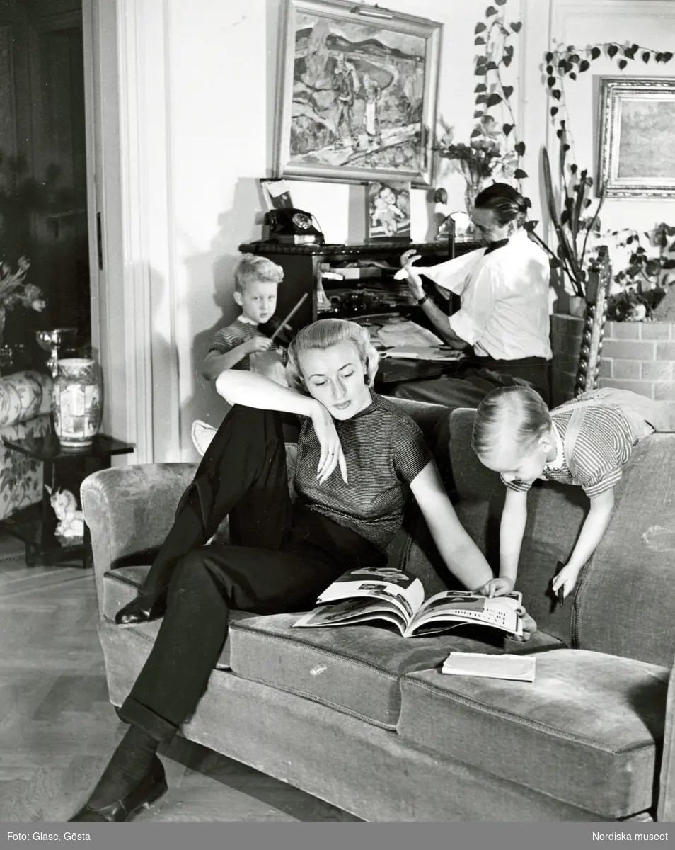 Kvinna, mannekängen Kim Söderlund, i randig t-shirt och byxor sitter i en soffa och bläddrar i bok tillsammans med pojke i hängselshorts. Man i vit skjorta sitter vid sekretär bredvid pojke med fiol.