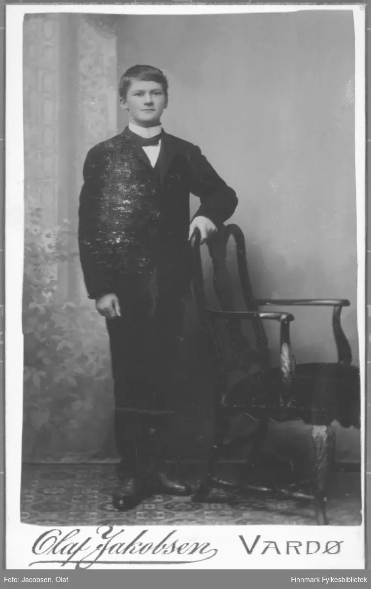 Portrett av en ung menn kledd i en mørk dress sløyfe og hvit skjorte med høy krage. studio ateliere malt bakgrunn holder hånd på stolrygg.  Albumet med bildet kommer fra Ekkerøy, kanskje han kommer derfra.