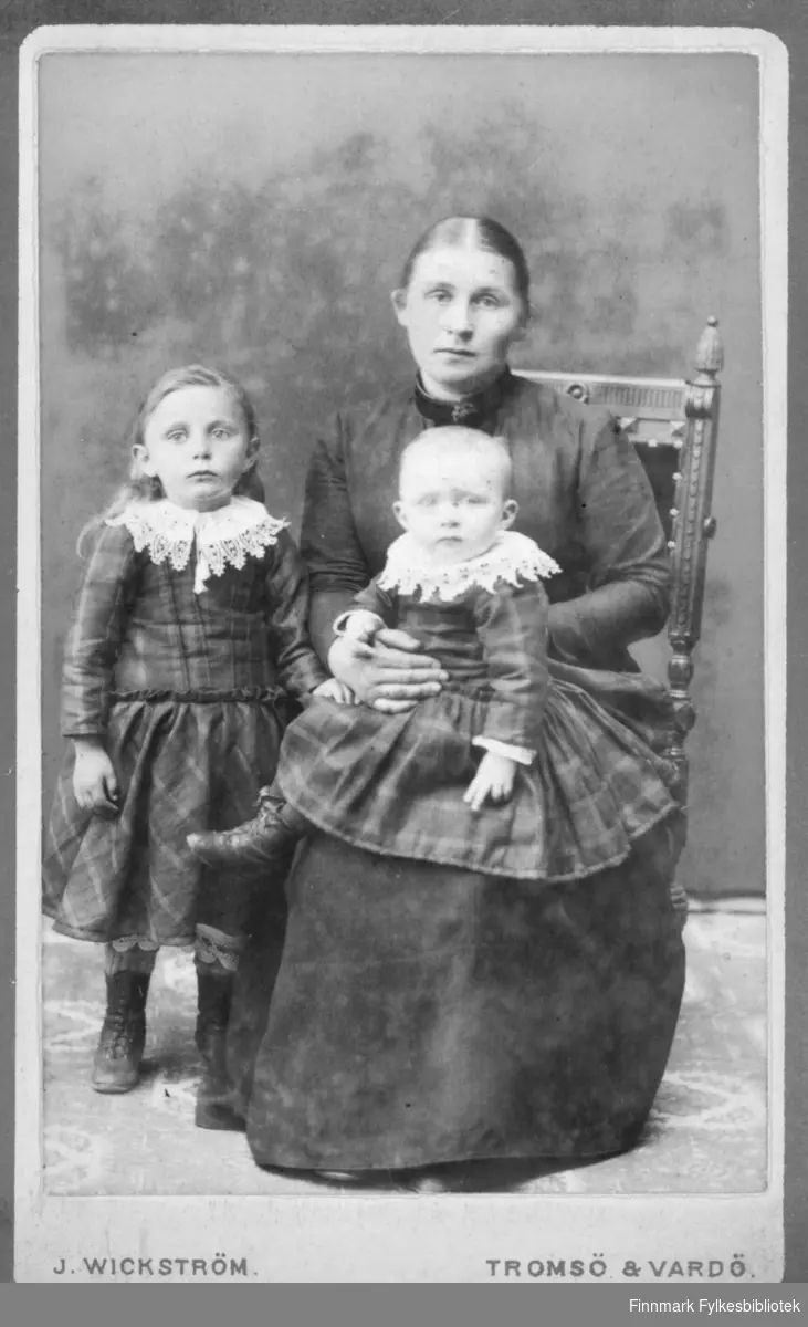 Familieportrett av en mor og hennes to barn tatt av Jørgen WikstrÃ¶m i Vardø. Kvinnen sitter med den minste jente i fanget, den andre jenten står ved sidan av henne. Jentene er kledd i like rutete kjoler med hvite broderte krager. Moren i en vanlig  mørk lang kjole. Albumet med bildet kommer fra Ekkerøy, kanskje familien bodde der.