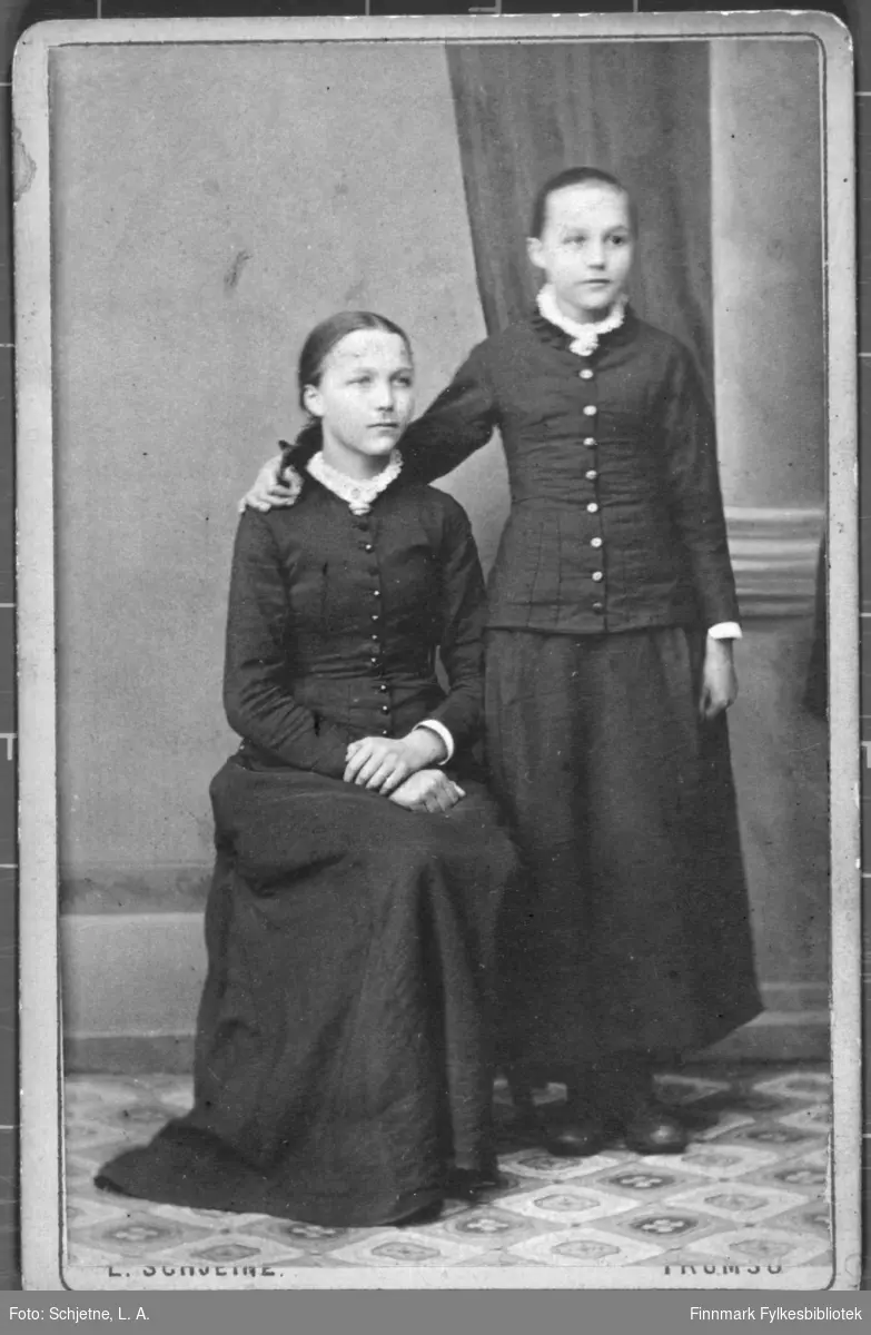 Familieportrett av to jenter i mørke drakter med hvite krager. studio ateliere. Jenta til høyre holder rundt skuldra på den andre. Albumet med bildet kommer fra Ekkerøy, kanskje de bodde der.