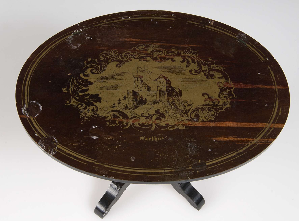 Divansbord av brunmålat trä med gulddekor. Motiv från Wartburg i guld på skivans ovansida. Bordskivan bärs upp av en pelare som delar sig i fyra fötter vid golvet.