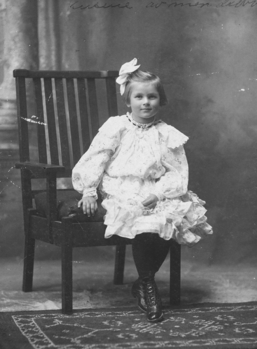 Et portrett av en liten pike. Hun er kledd i en vakker kjole og har på seg strømper og sko. I håret har hun en hårsløyfe og et perlesmykke rundt halsen. Hun sitter på en stol. På gulvet foran henne kan man se en matte.  Bildet trolig tatt på begynnelsen av 1900-tallet.