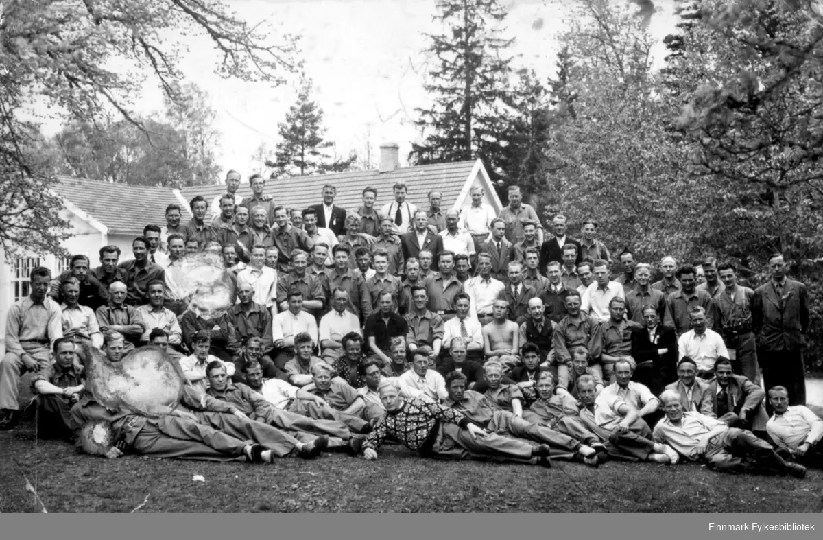 Fotografi av en stor gruppe menn som er hentet hjem med de hvite bussene ifra tysk fangenskap under andre verdenskrig. Magnus Stenbakk sitter i tredje rekke nedenfra, omtrent midt på bildet. Han er kledt i en mørk skjorte. Bildet er tatt ute på en gressplen. I bakgrunnen ses en del av en hvit bygning