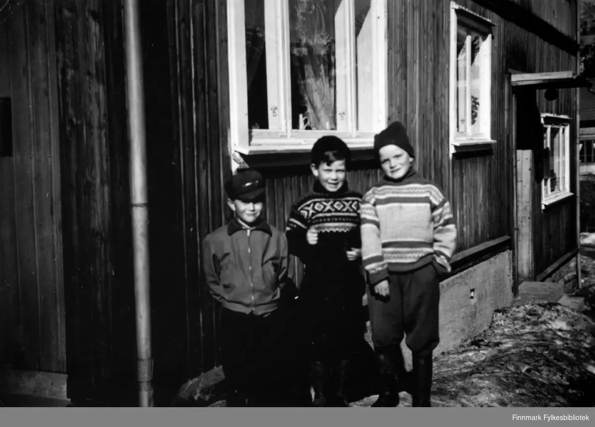 Fotografi av tre ukjente gutter som står utenfor et trehus. Gutten i midten har på seg Mariusgenser.