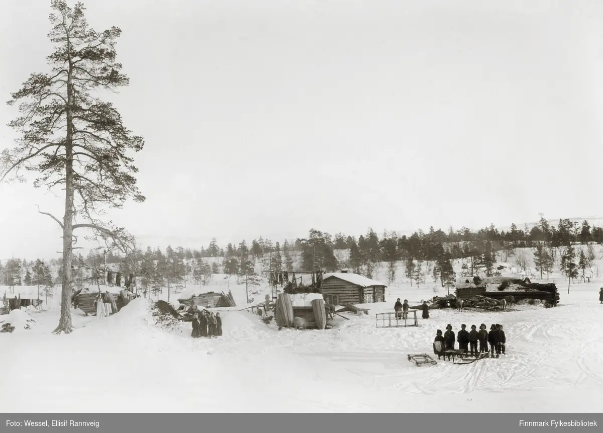 Tømmerhus og hytter i Øvre skolteby fotografert om vinteren. En gruppe menn står samlet om en slede i høyre hjørne av bildet nærmest kamera, en gruppe kvinner et stykke lenger til venstre
