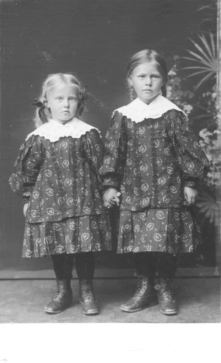 Portrett av to jenter, ikke navngitt, antakelig søstre siden de har like kjoler. Kjolene er mønstrete med hvite blodekraver, begge jentene har flettet hår. På føttene har de snørestøvler. Fotografert i fotostudio med en malt bakgrunn med palmer. Den minste jenta kan være ca. 5 år og den eldste ca. 7-8 år.