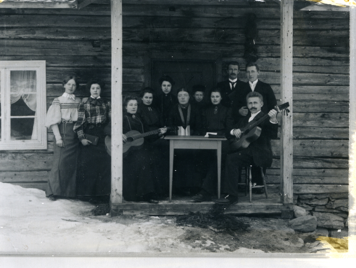 Kvinner og menn oppstilt framfor inngang på tømmerbygning, to med gitarar. Mannen med gitar til høyre i bildet er emmisær Hans Evensen.