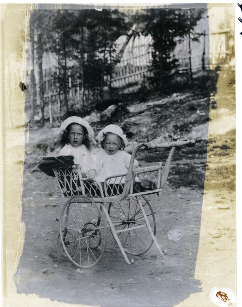 To jenter sittende i barnevogn . Sigrid f. 1915 gift Hall og Synnøve f.1916 gift Kolstad. Bildet er tatt utenfor kjøpmann Melby sin forretning og jentene er hans døtre. Bildet er tatt ca 1917.