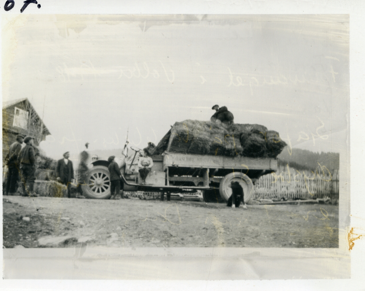Lastebil med forlass foran stakittgjerde. Tømmerbygning til venstre. Barn, karer og en hund er med på bildet. Jørgen Dale er sjåfør 
(1897-1979).Bildet er tatt rundt 1918-1920