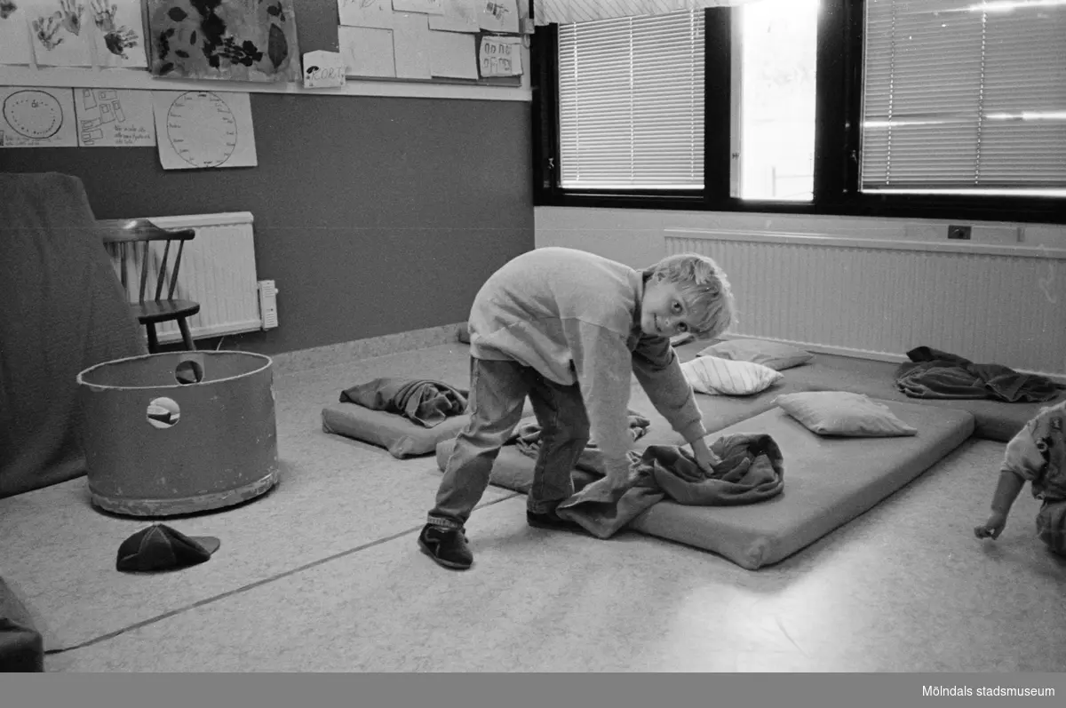 En pojke tittar leendes mot fotografen. Han lutar sig ned och håller på en filt som ligger på en madrass på golvet. På madrassen ligger en kudde. Kanske dags för en liten sovstund? Katrinebergs daghem, 1992.