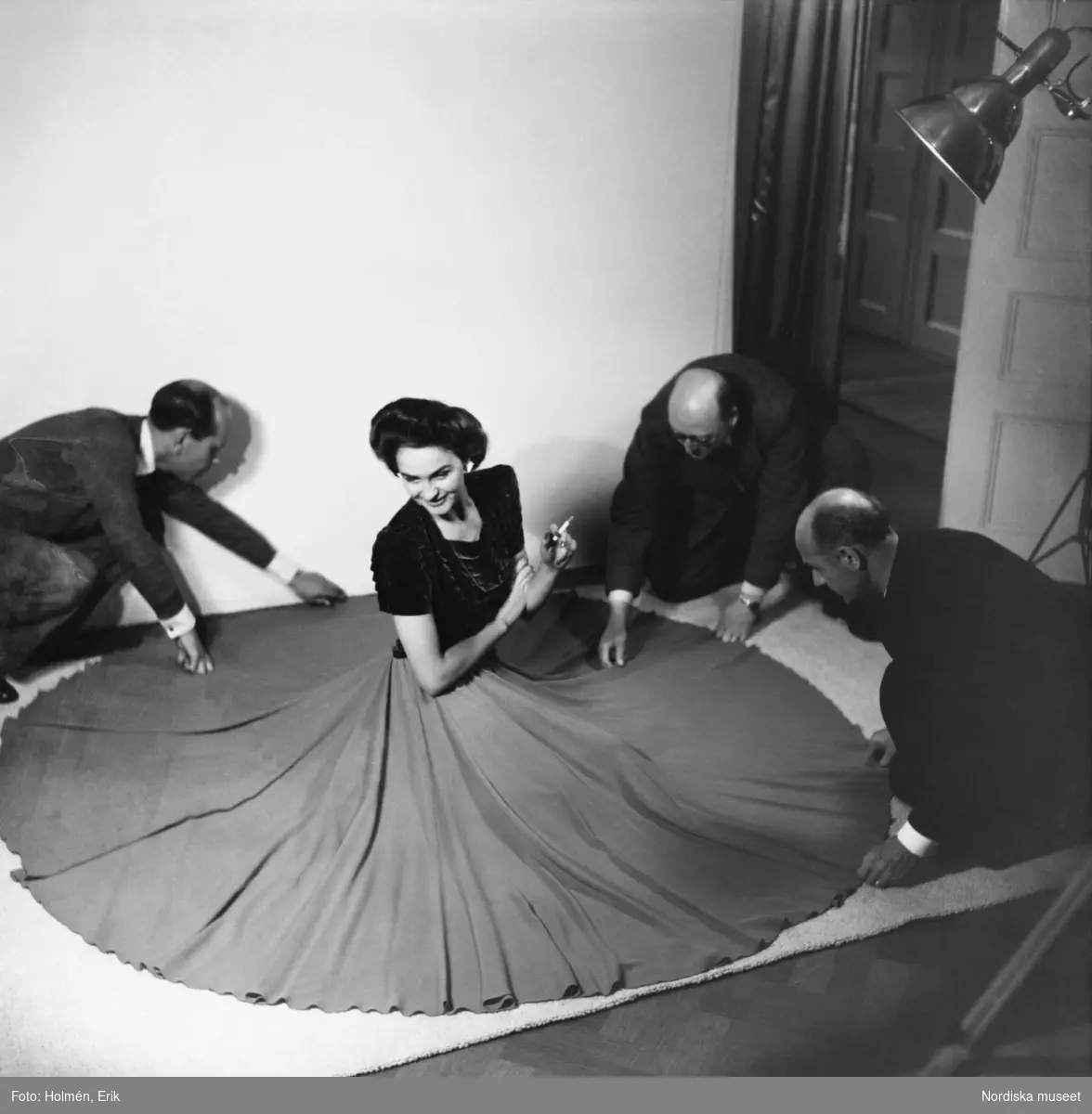 Modellen Hjördis Genberg fotograferas för Stil. Hon sitter på en vit matta, klädd i mörk överdel och vid kjol med en cigarett i handen. Tre män, fr. v. herr Paulsen, herr Hilgardt och kapten Ungervitter, draperar kjolen i en cirkel på golvet.