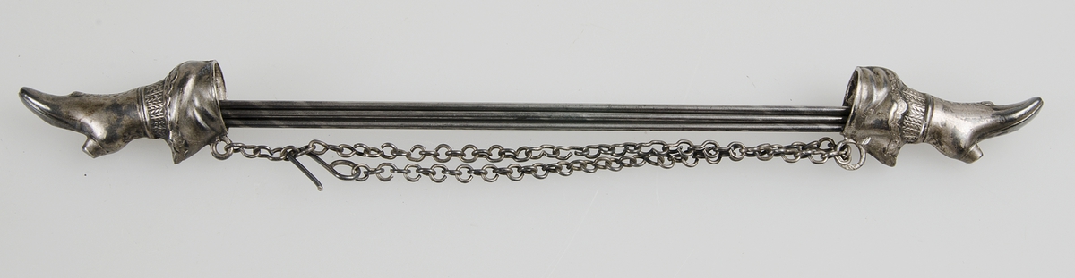 Fem strumpstickor av stål i strumpstickshållare i form av två kängor av vitmetall med kedja som håller samman stickorna.