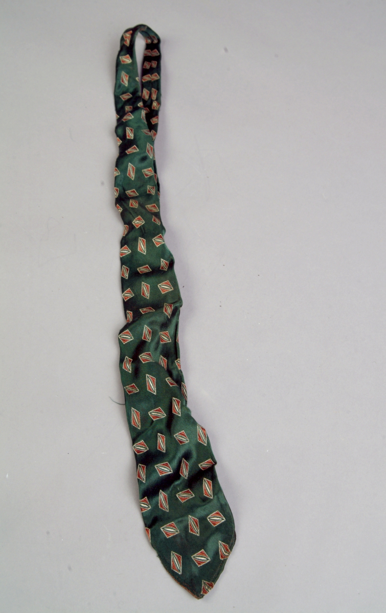 Et langt og relativt smalt slips sydd av silke (sateng). Bunnfargen er mørk grønn og det er ivevd dekor i form av ulike firkanter i rustrødt og beige. Slipset er maskinsydd. Det er avrundet spisst i begge ender.
