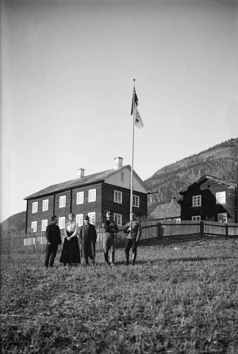 Fire menn og en kvinne, i bakgrunnen et hus og et flagg