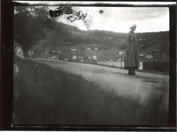 Kvinne alene på en vei. Utsikt over et vann, hvor flere hus 