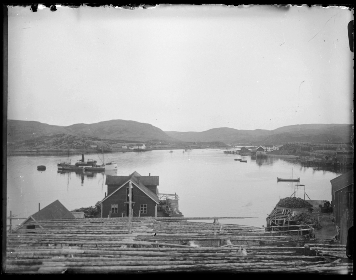 Båtsfjord havn, oversiktsbilde. Til venstre et mindre dampskip under lasting/lossing - dette kan være enten et postskip eller en lokalbåt. Vi ser tomme fiskehjeller, kaier, pakkhus og andre bygninger. Fiskebruket i front tilhørte J. E. Abrahamsen.