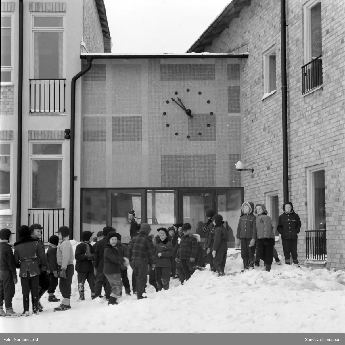 Mosaikvägg med klocka ovanför en av entréerna på Sankt Olofsskolan. På skolgården framför leker barnen i snön.