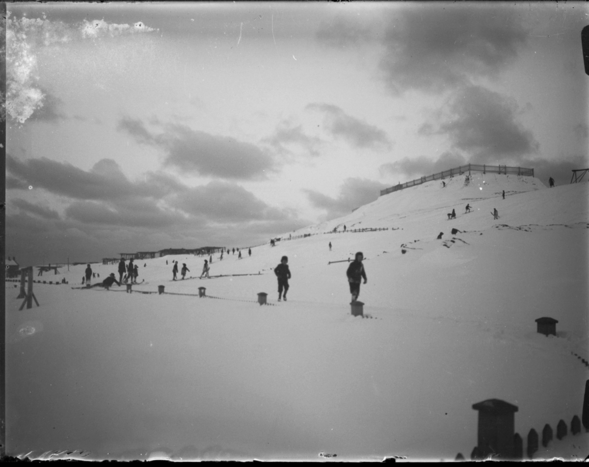Vinterbilde fra Vardø. Barn, noen med ski og kjelker, leker i området mellom prestegårdshagen og Klondyke. De bruker Klondykebakken som akebakke og skibakke