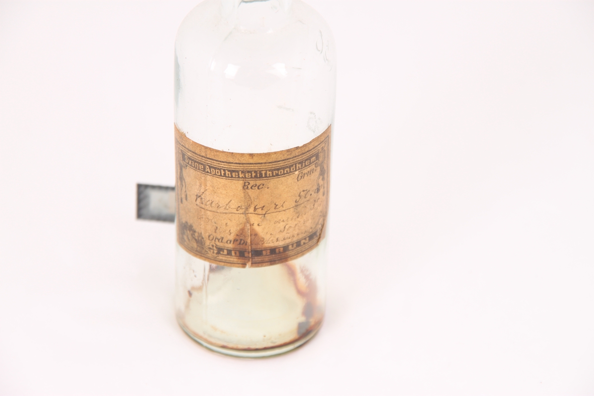 Liten glassflaske med kork til oppbevaring av medisin. Flasken inneholder spor etter tørket væske, muligvis karbolsyre iflg. etiketten.