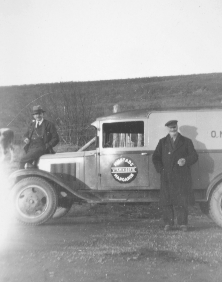 To menn ved siden av en bil som kan være en Chevrolet varevogn, 1930-33 mod. En står ved siden av bilen, og den andre sitter på panseret. Det er et stort rundt merke på døra til bilen. Det står Mustad margarin.