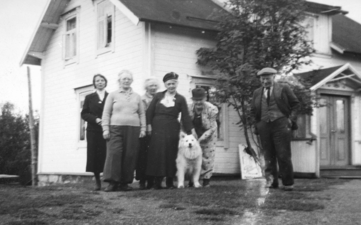 Fem damer og en mann + en hund står foran en bygning.