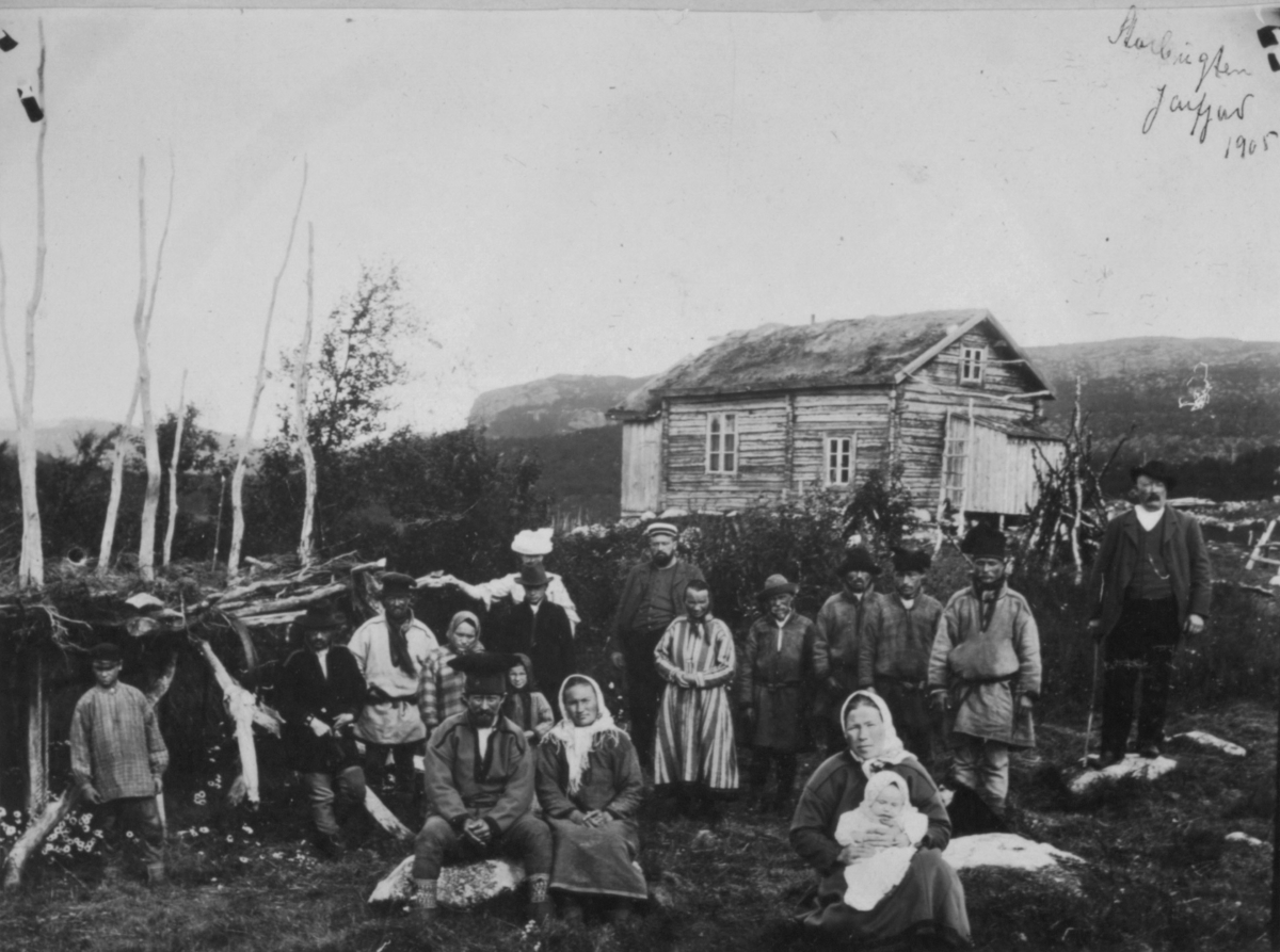 En gruppe menn go kvinne i samiske drakter fotografert i Jarfjord i 1905. Fremst på bildet en kvinne med et spedbarn i fanget. Bakerst står det et tømmerhus.