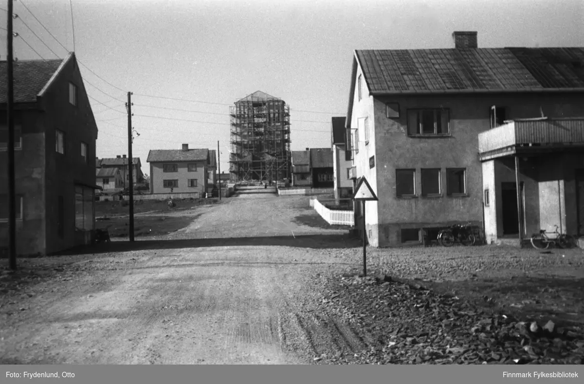 Kirkegata i Vadsø fotografert nordover, opp mot kirka som er under bygging i 1957 (kirka ble vigslet 30. mars 1958). Til venstre nærmest kamera Bjercks forretning i sentrum, til høyre Mathisens butikk. Lenger oppover gata ser vi gjenreiste boliger.