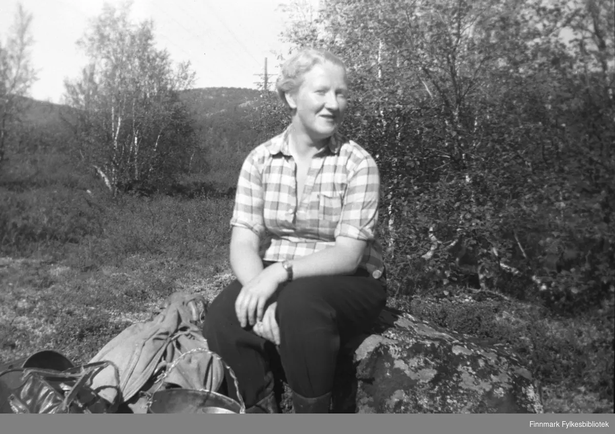 Herlaug Mikkola på bærtur i Neiden-distriktet. Hun sitter på en stein, og foran henn ser vi ryggsekk og bærspann