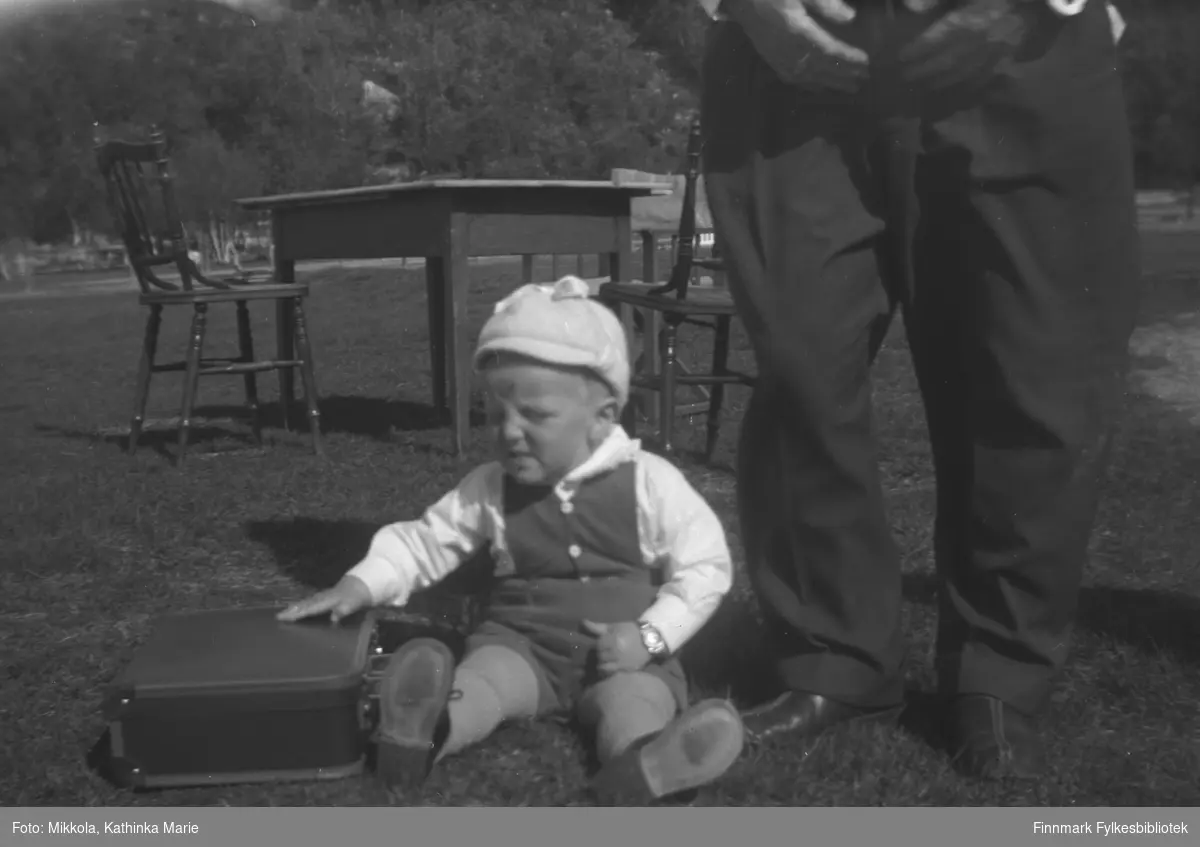 Asle Lindseth leker med en koffert i gresset utenfor Mikkelsnes. Til høyre i bildet ser vi beina til bestefaren, Aksel Konrad Mikkola