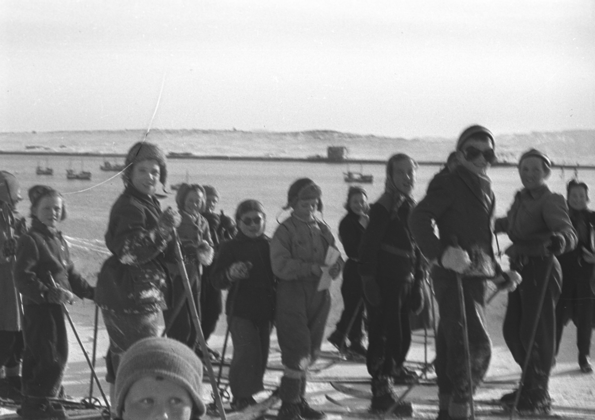 Mange barn deltar på skirenn i Vadsøs gater på 50-tallet. Havna med flere båter fortøyd ses i bakgrunnen. Personene er ukjent.