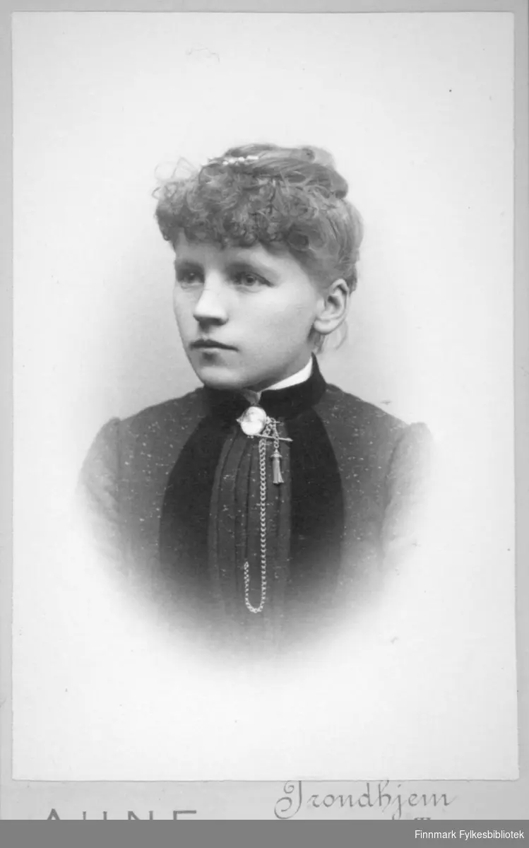 Portrett av en dame iført en mørk overdel. En liten flik av den hvite skjorta/blusen hun har under vises i halsen. Der ses også et langt kjede.
