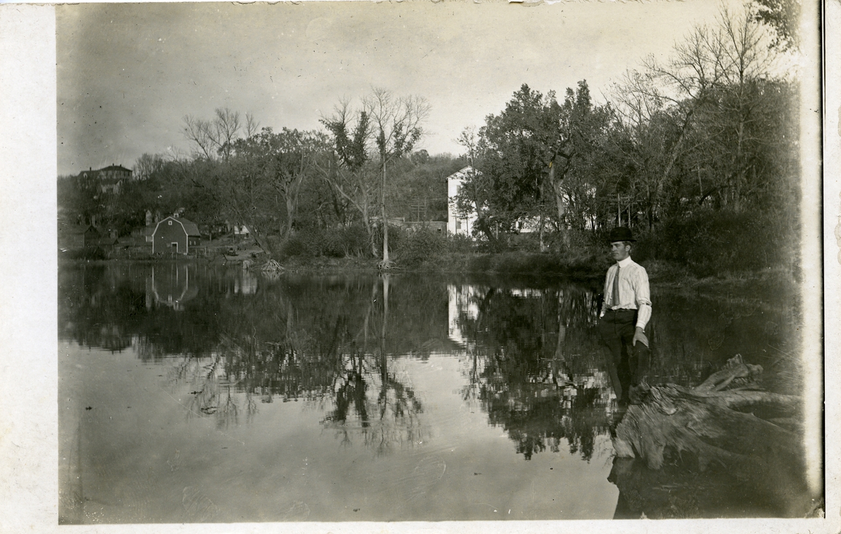 Mann stående på ei trerot i et vann/innsjø. Hus i bakgrunnen. Dette er et postkort
1911-1912