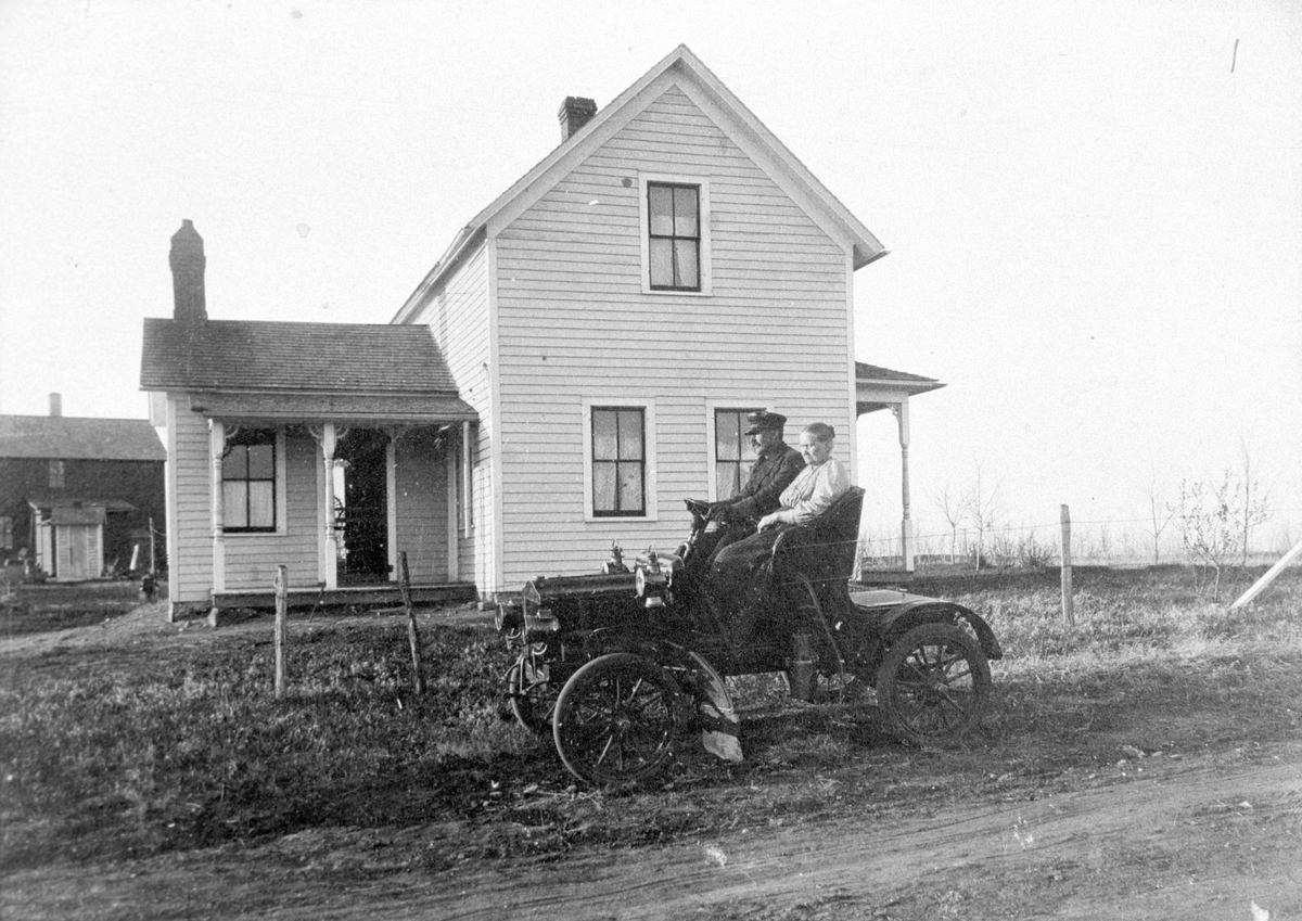 Amerikabilder, Per Mæhlum, Vallset.
Bil med to passasjerer, bolighus i bakgrunnen, Eastedge, Nord-Dakota, USA. Mannens navn er Remmen, og kvinnen ved siden av er hans mor. Disse to var fra Stange. 
Fotograf er Per Mæhlum, ca 1910.