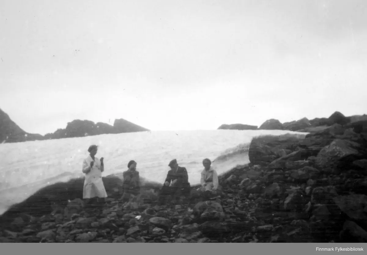 Fire personer sitter ved fjæra. Snøen er ikke helt smeltet, vi ser rester av vinterens snø i bakgrunnen. Damen til venstre på bildet har en lys kjole med korte ermer på seg. Ved siden av henne sitter en dame i ganske mørke klær. Mannen har en mørk dress og sixpence på hodet. Damen til høyre på bildet har et lyst skjørt på seg. Stedet er i Bergsfjord i Loppa rundt 1920-1930.