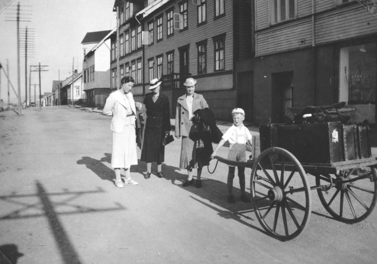 Tor Hauge står og holder skjekene på ei handvogn, med store hjul. Kvinnen i hvitt er hans mor Frida Hauge. De to andre damene er besøkende fra Bergen. Bildet er tatt i Havnegata