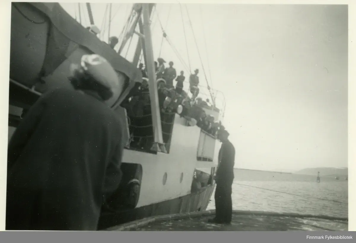 Medlemmer av Vadsø damekor ombord et skip, muligens hurtigruten. Flere av damene har på seg hvite hatter (trolig kor uniform) og kåper. På kaia kan man se en mann og en kvinne. På siden av skipet kan man se at det henger en livbåt.