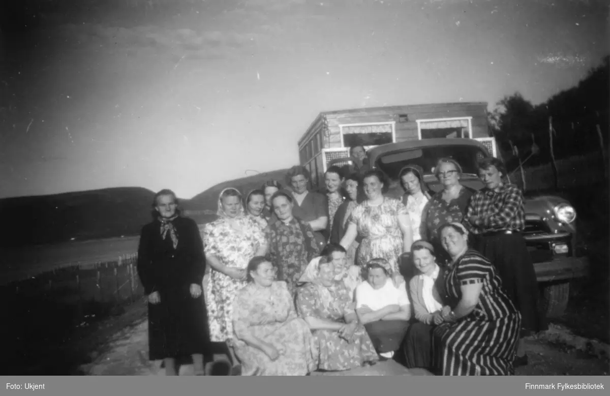 Nesseby husmorslag er ute å reiser. Her poserer de foran en lastebil Chevrolet 1954 med campingvogn. Kvinnene har på seg kjoler, skjørt og skjorter. Enkelte har på seg skaut, andre har briller. På campingvognen kan man se et par vindu med gardiner i. De står parkert på kanten av en vei. I bakgrunnen kan man se fjell.