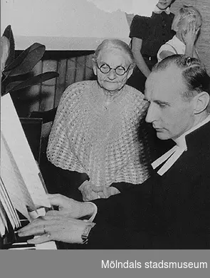 En präst spelar piano och en äldre kvinna sitter bredvid och lyssnar. Okända personer.