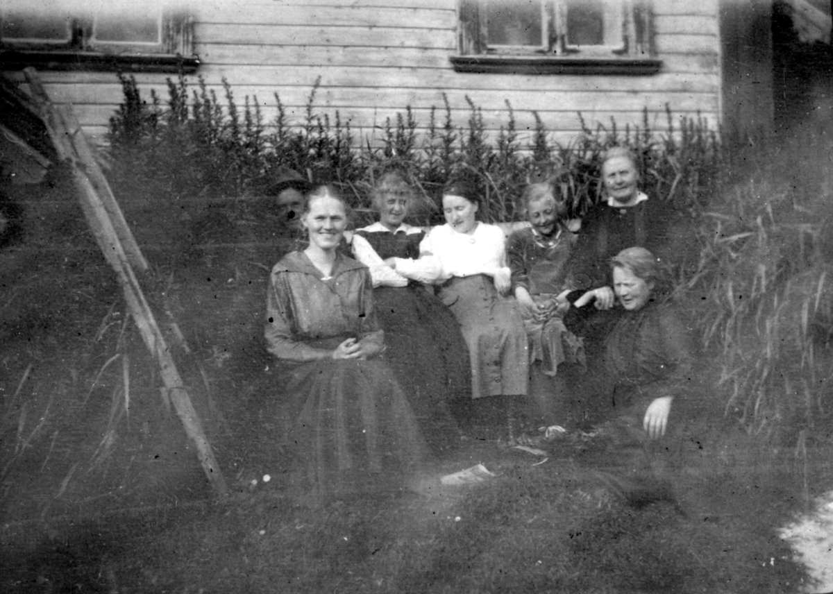"Hos kjøpmann Buch. Hasvik." Muligens familien- Buch som poserer for et bilde. Flere kvinner sitter på en benk kledd i kjoler. En mann sitter bak benken. Gruppen er omringet av planter, og bak gruppen kan man se et trehus.