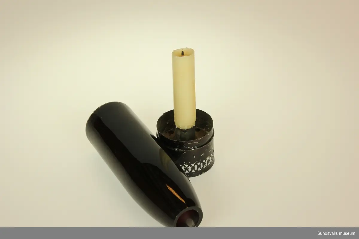 Mörkrumsbelysning bestående av en svartmålad hållare för stearinljus samt en avlång kupa i röttglas.
