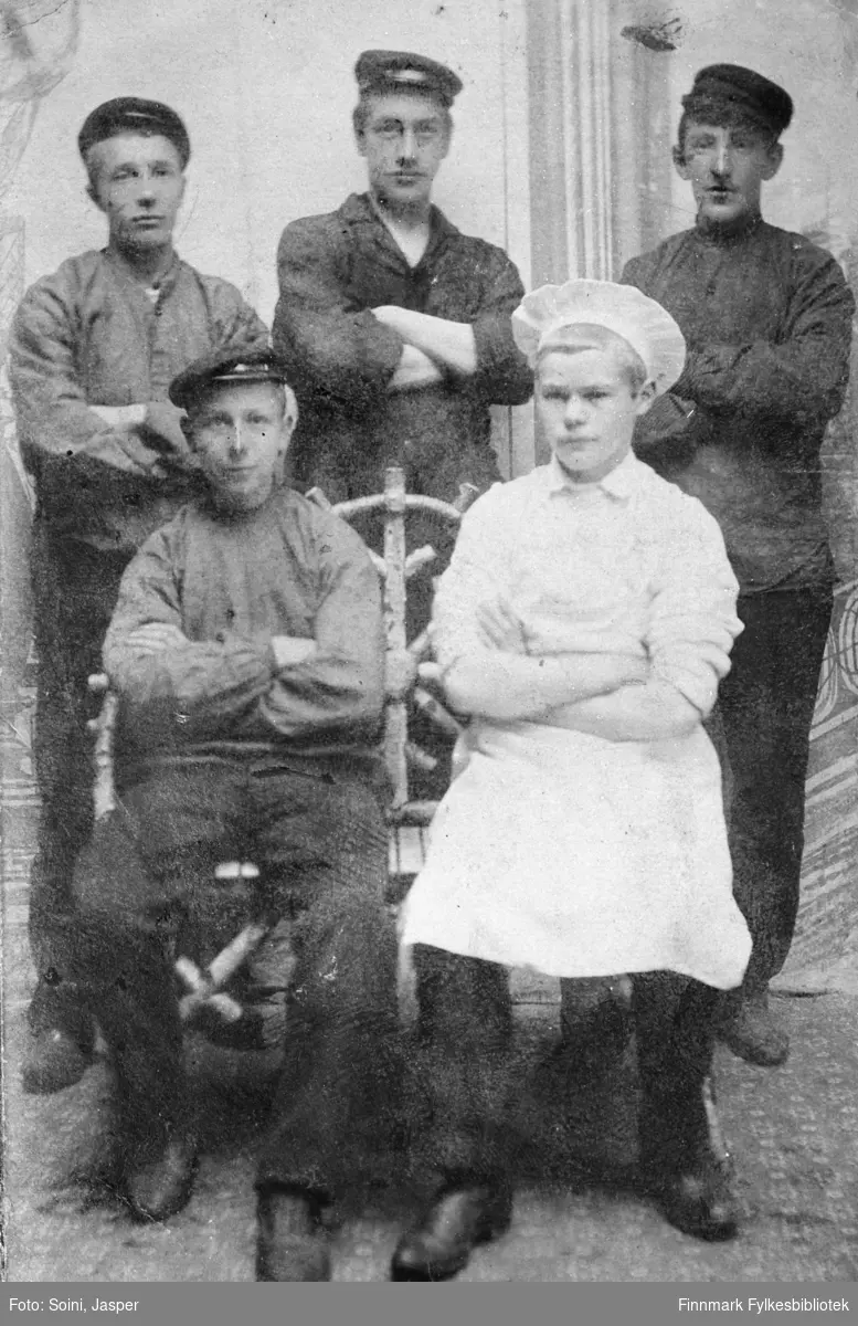 Gruppebilde av fem unge menn tatt hos fotografen Soini i Vadsø. Georg Bjerk og Edvard Pettersen sittende foran, den sistnevnte kledd som kokk, og J.Korbi, Eivind Riise og A.Hågensen stående bak dem. Bildet er tatt ca. 1905-1910.