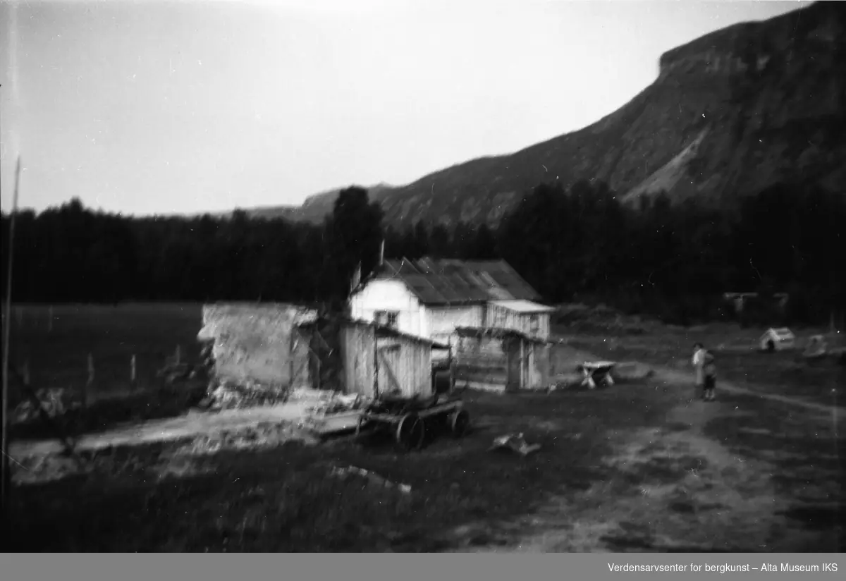 Oversiktsbilde som viser tunet på Jøraholmen, med et lite hus og noen ruiner. To personer til høyre på bildet.