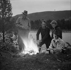 Fire personer sitter rundt et bål på elvebredden og drikker 