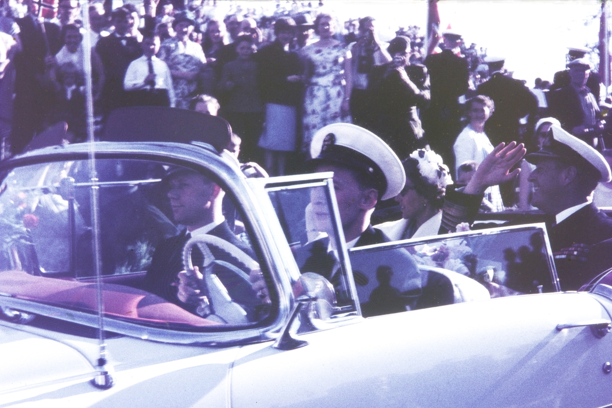 Kongen hilser fra bil. Folkemengde i bakgrunnen.
