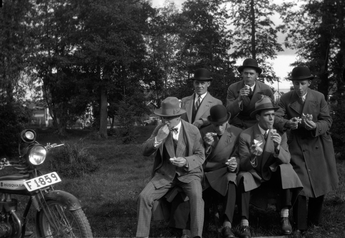Några män från föreningen Tord tar matpaus och de äter kokt korv i papper och bröd i andra handen. Till höger sitter Einar Svensson och i mitten upptill står "Nisse Falköping". En Husqvarna motorcykel står parkerad bredvid dem.