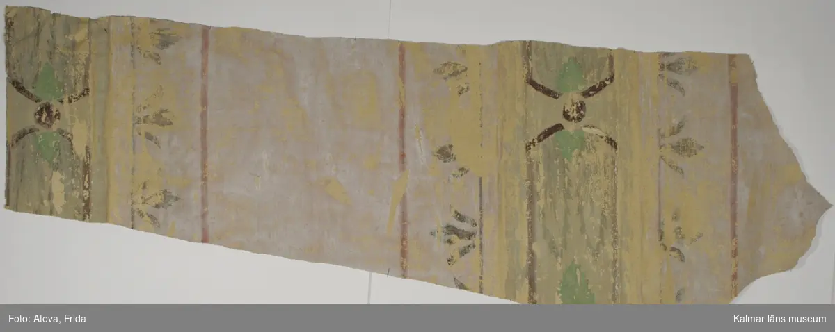 KLM 28335:2. Tapet av papper. 2 st bitar. På ena sidan helt vitmålade. På andra sidan syns avtryck av en schablonmålning. På mitten syns avtryck av KLM 28335:3. Tapeten har alltså suttit över :3. Schabloner med stiliserade ornament i lila, grönt, brunt och rosa. Datering: 1800-tal.