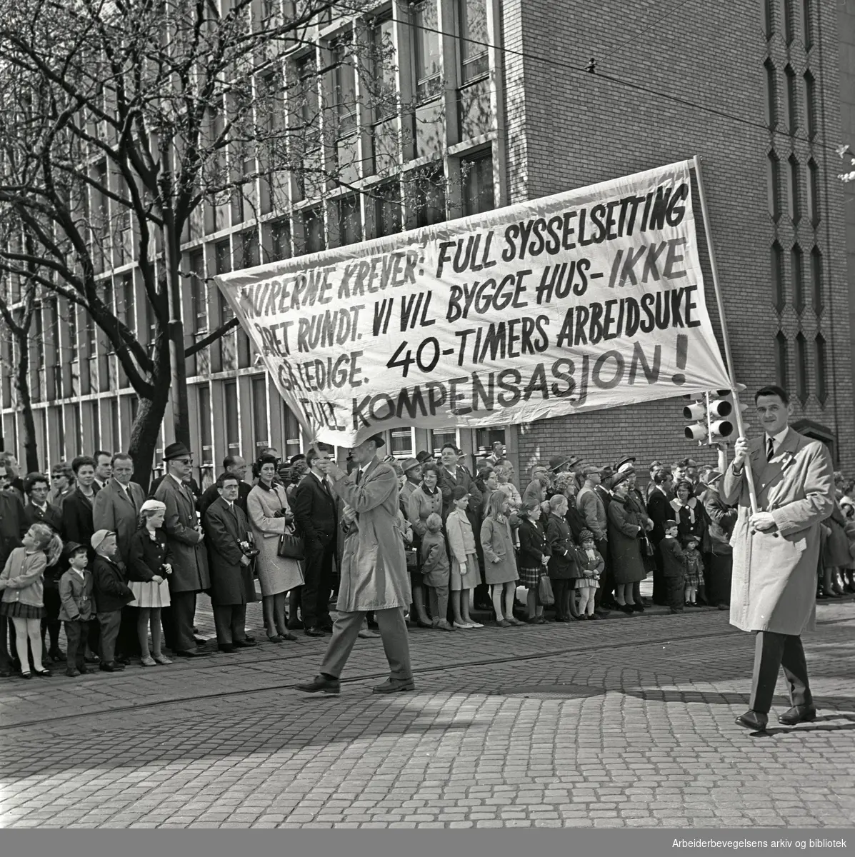 1. mai 1964 i Oslo.Demonstrasjonstoget i Karl Johans gate.Parole: Murerne krever: Full sysselsetting året rundt. Vi vil bygge hus - ikke gå ledige. 40-timer arbeidsuke. Full kompensasjon!.